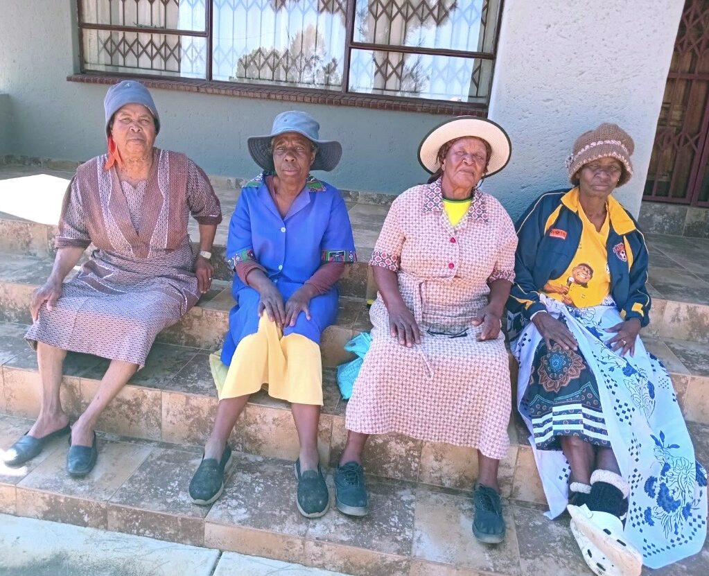 Four seniors sitting together on the steps outside a home in Ga-Kgole, South Africa, representing the communities supported by Love and Light Wellness. Copy